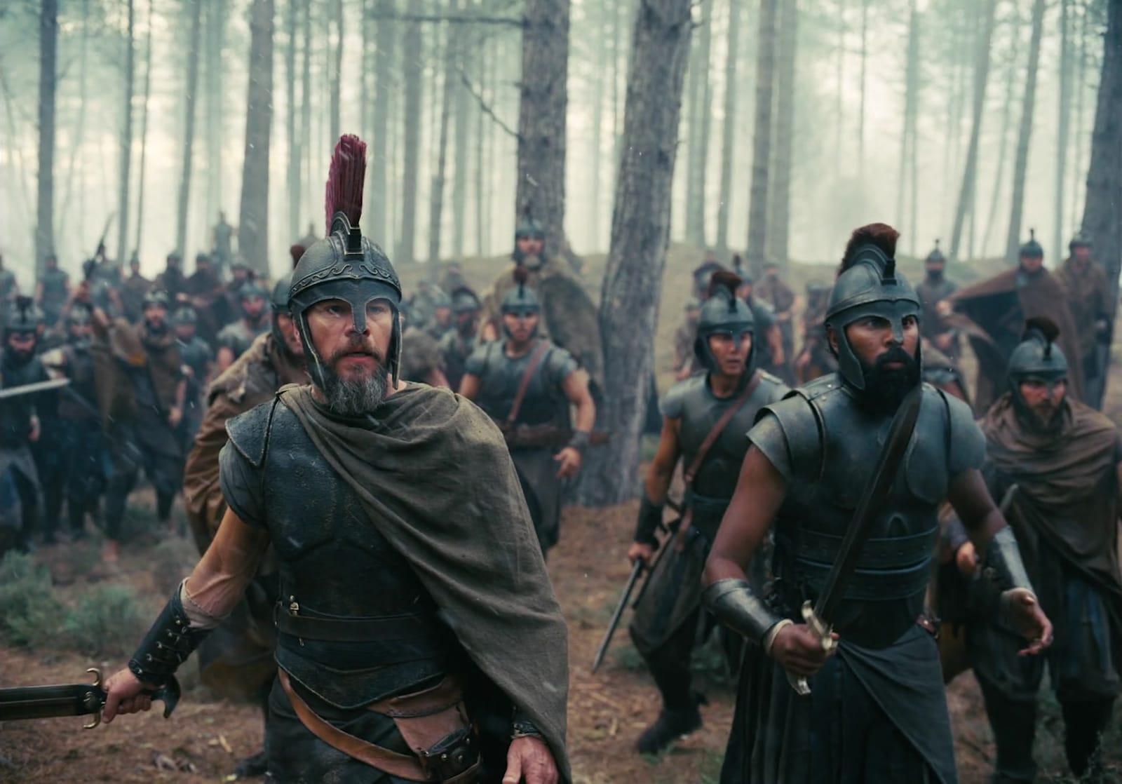 Odysseus, in a red-crested helmet and a green cloak, sword low in his hand, leads his armed men through a misty pine forest; the full frame holds the treetops above them and the forest floor below.